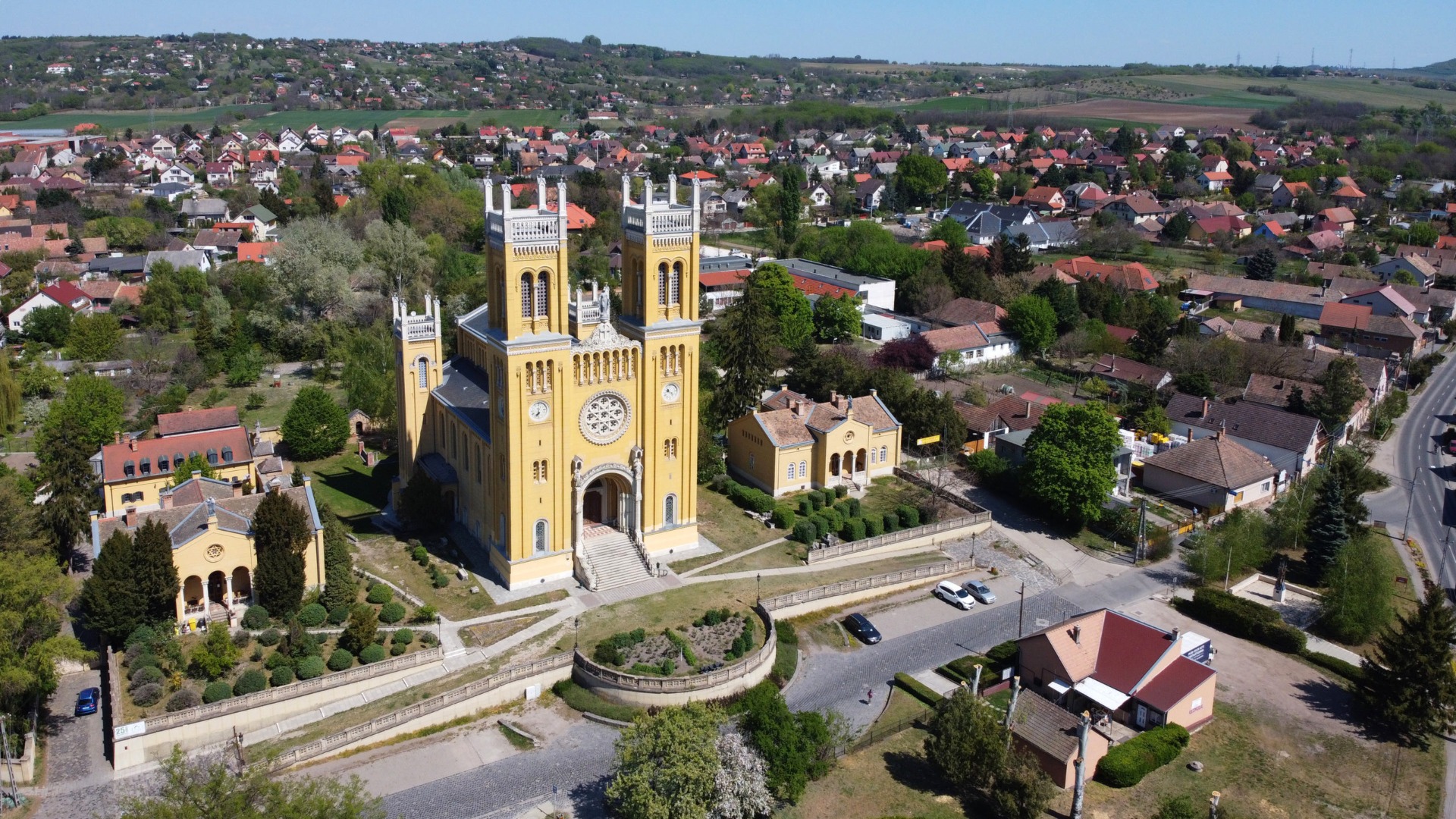 Fóti sárga tornyos templom légifelvétele, körülötte park és házak, dombvidéki városrész a háttérben.
