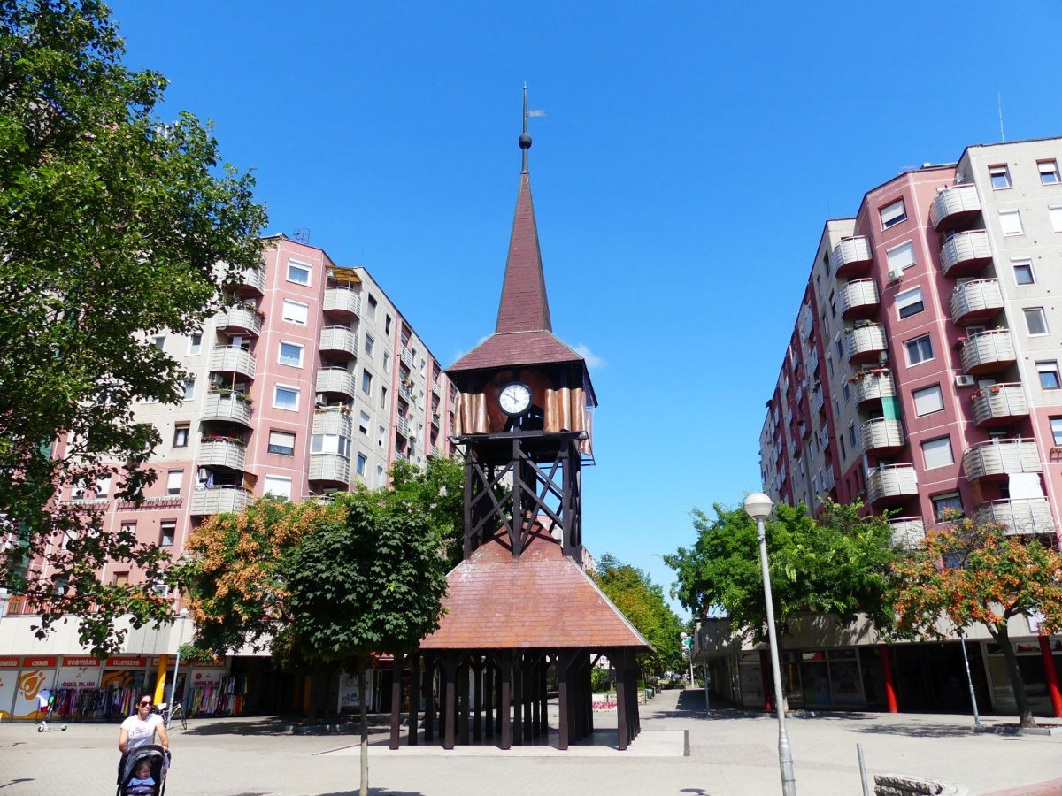 Káposztásmegyer városi tér közepén fa harangtorony órával, körülötte magas lakóházak és zöld fák.